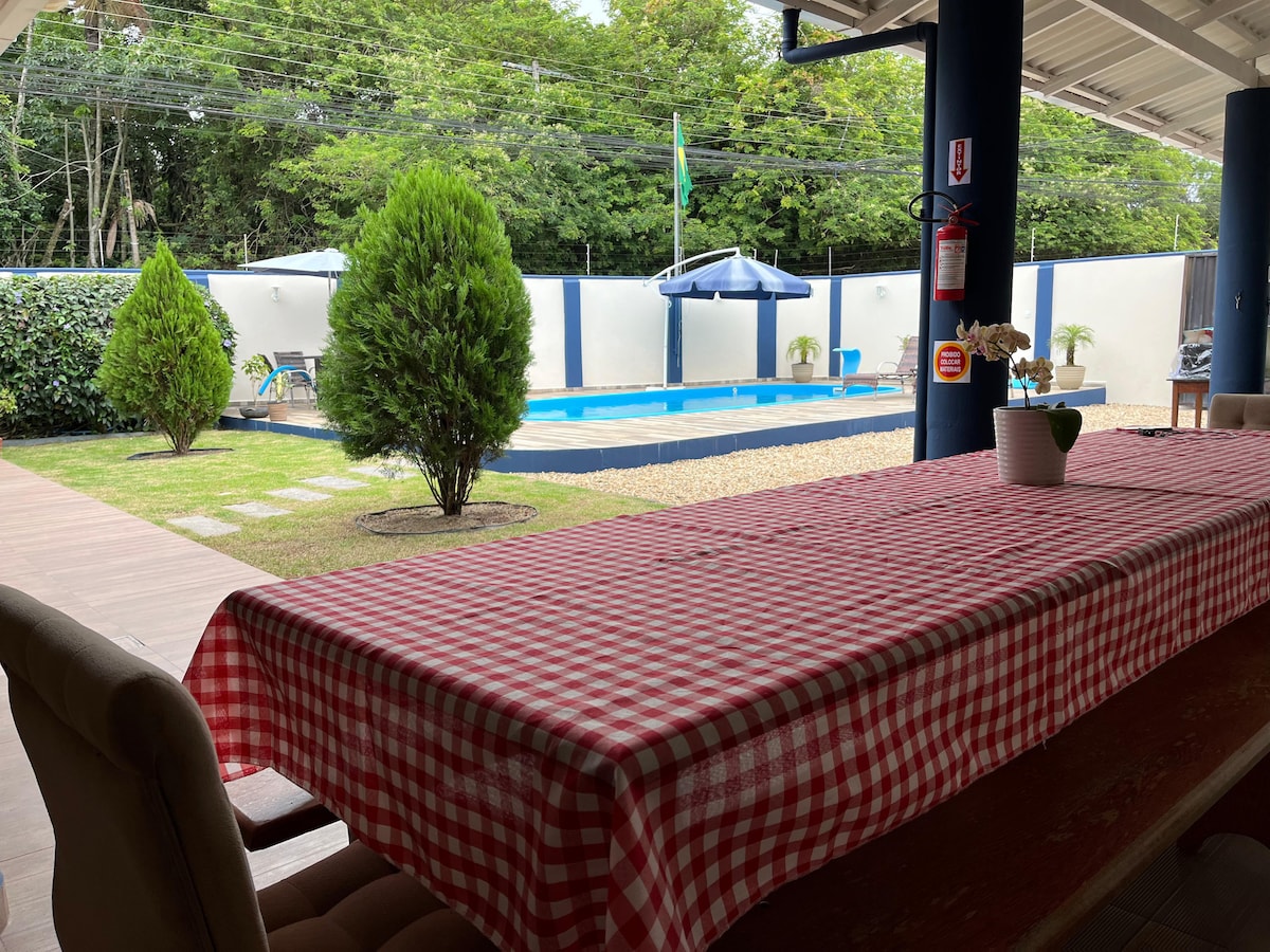 A spacious outdoor dining area is presented with a checkered tablecloth, facing a well-maintained pool surrounded by green landscaping. A shaded gazebo is visible in the distance, offering a comfortable spot for relaxation.
