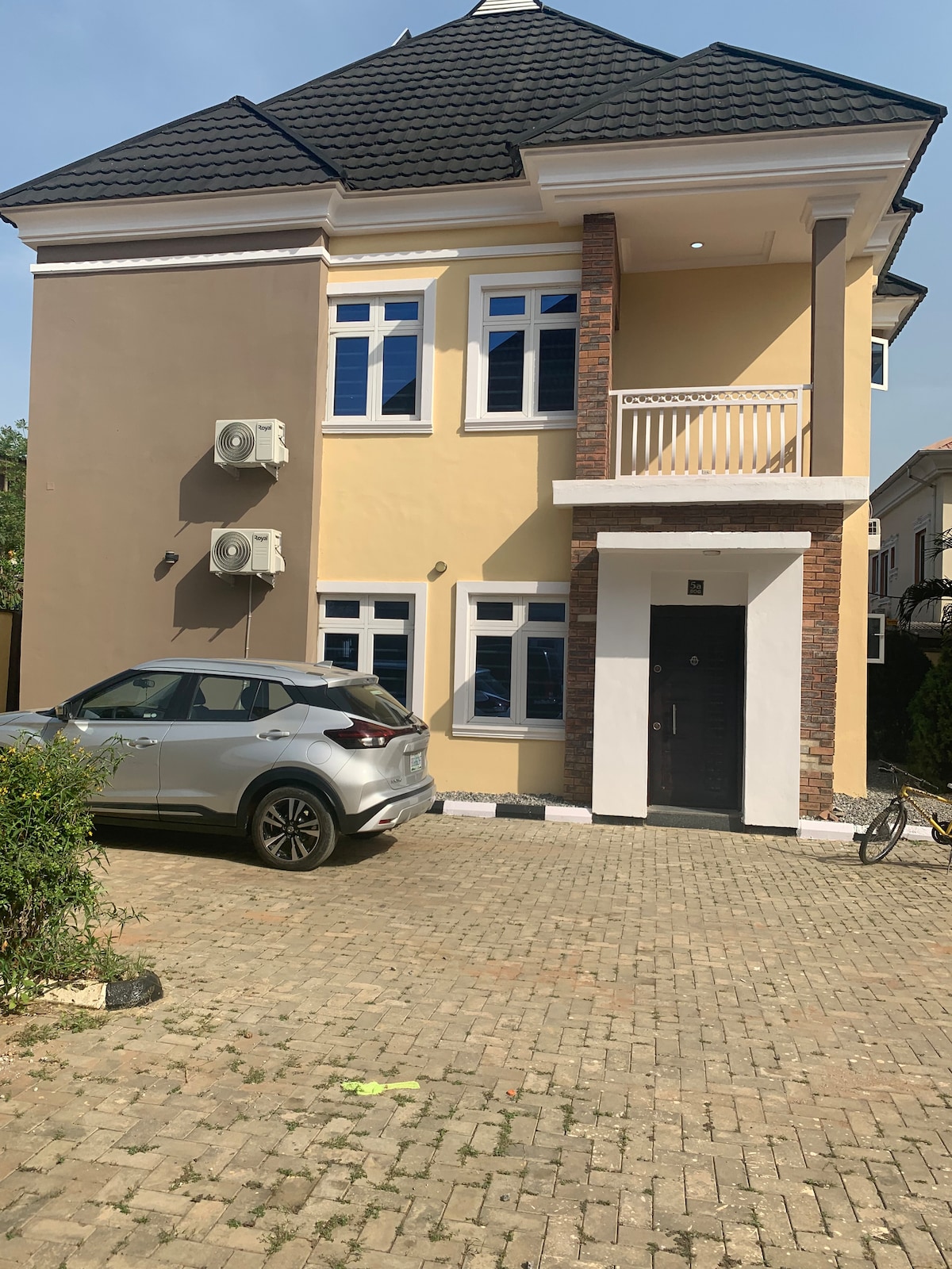 The exterior of the two-story house displays a modern design with beige walls and a stylish roof. Large windows allow ample natural light, while two air conditioning units are installed on the side. A paved driveway leads to the entrance, featuring an automated keyless entry.