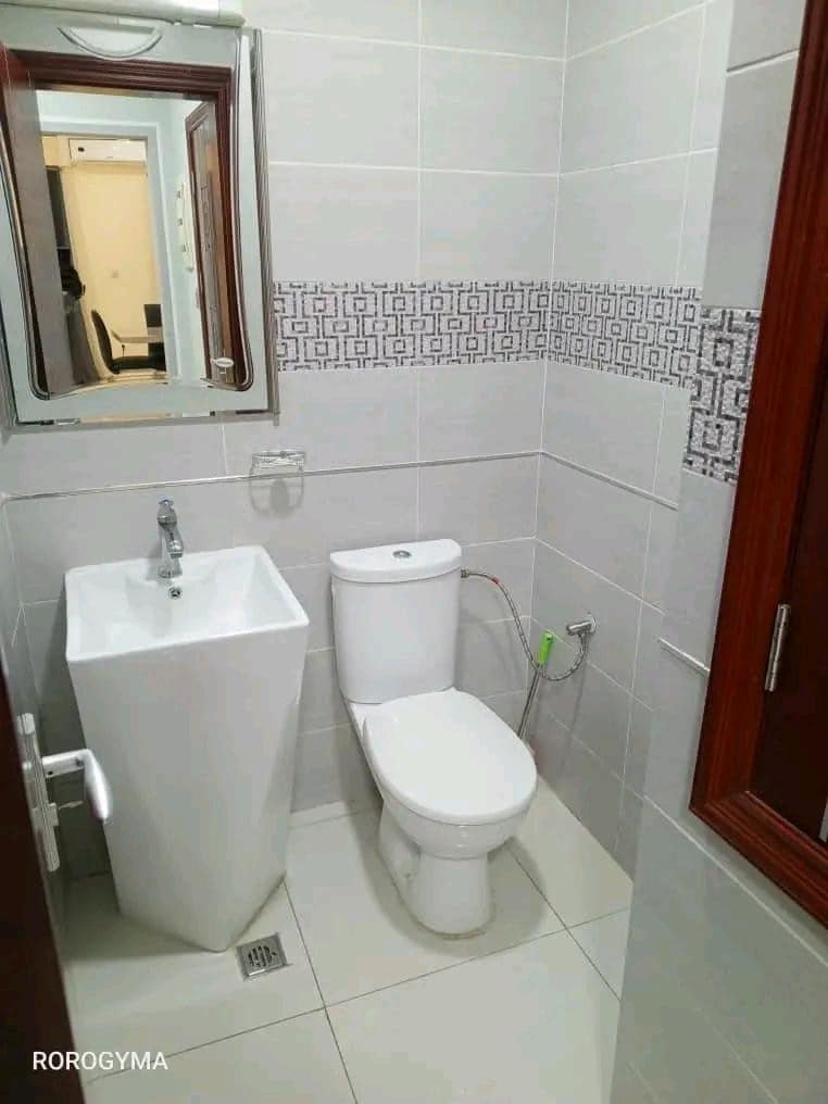 A modern bathroom is designed with sleek tiles and a minimalist aesthetic. A tall, rectangular sink is positioned next to a white toilet. A large mirror above the sink reflects the clean, organized space, while subtle patterned tiles add visual interest to the wall.