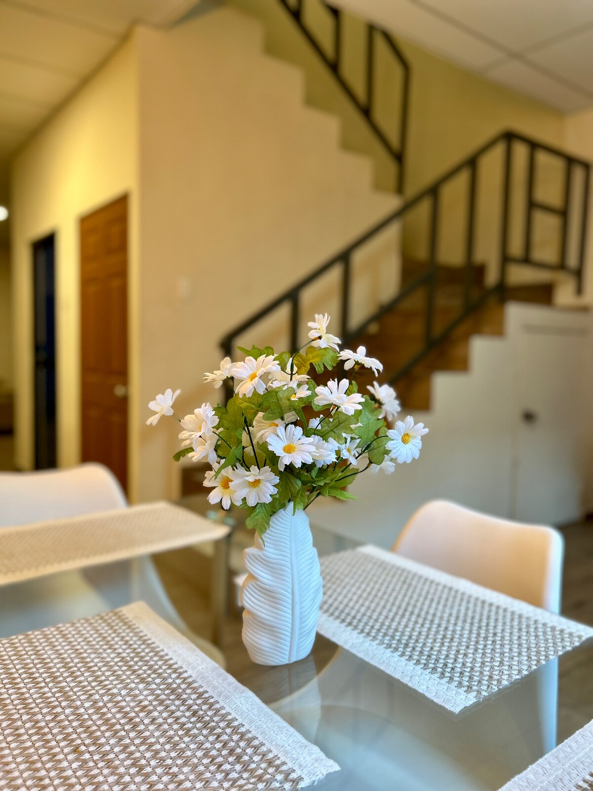 A dining area is presented with a glass-topped table adorned by a vase filled with white daisies. Light-colored placemats complement the table setting. In the background, a staircase is visible, providing access to the upper level of the home.