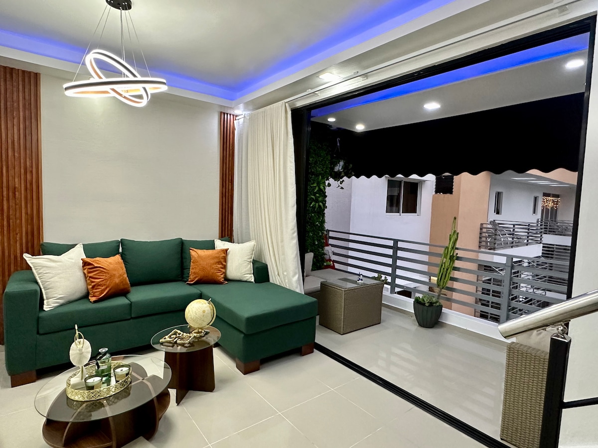 A stylish living area features a green sectional sofa adorned with decorative pillows. A modern light fixture hangs overhead, illuminating the space. Glass doors open to a balcony with potted plants, enhancing the outdoor connection. A coffee table with decorative accents is positioned in front of the sofa.