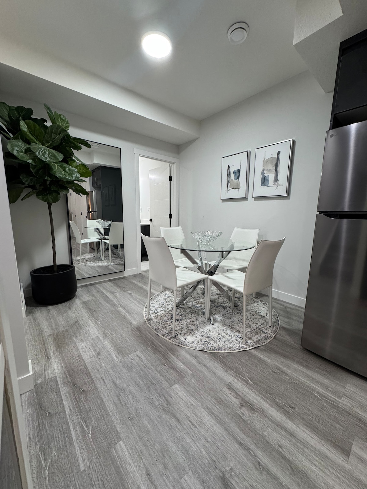 A dining area features a round glass table surrounded by four white chairs, situated on a soft, circular rug. A large potted plant adds a touch of greenery, while elegant wall art complements the neutral-toned walls. A large mirror reflects the space, enhancing brightness.