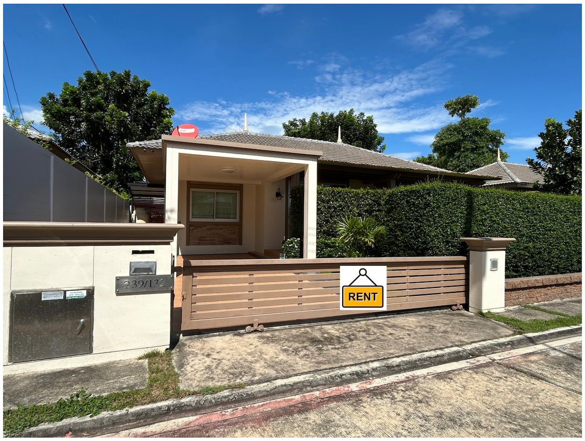 A single-story house is visible behind a wooden fence with a 'RENT' sign prominently displayed. Lush greenery surrounding the property offers privacy, and a clear sky is seen above, enhancing the inviting appearance of the home.