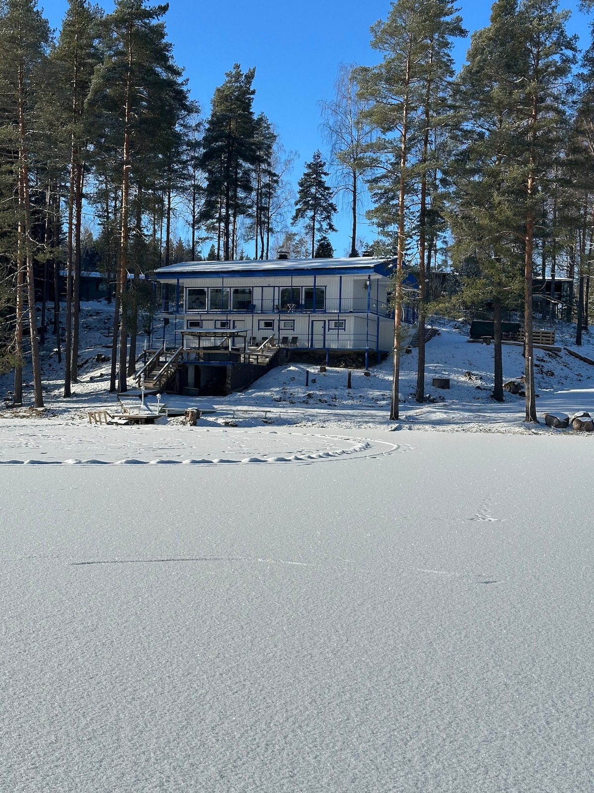 A contemporary two-story villa is set against a backdrop of tall pine trees. The building features large windows, and a sunlit deck extends towards a snowy landscape, reflecting a serene winter atmosphere. The frozen lake in front is blanketed in white, creating a peaceful setting.