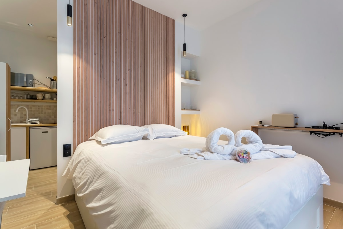 A spacious bedroom features a large double bed with crisp white linens. Decorative towels are arranged on the bed, and warm lighting illuminates the space. A wooden panel accent wall adds texture, while shelves hold neatly arranged items, contributing to a calm ambiance.