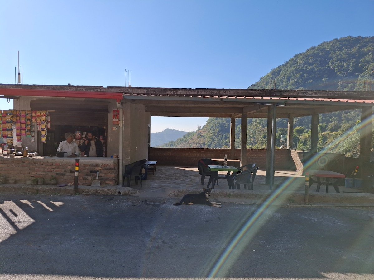 A rustic outdoor dining area is featured, with several black plastic chairs positioned around wooden tables. A small local establishment is visible in the background, where people are seen engaging with customers. A dog rests on the ground near the entrance, with a mountainous landscape in the background.