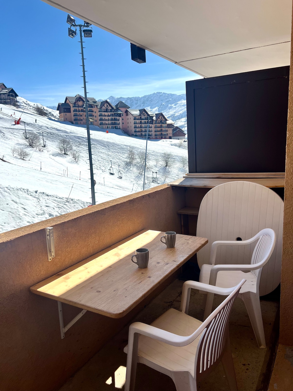 A small balcony features a wooden table with two cups, complemented by two white plastic chairs. The backdrop displays snow-covered slopes and buildings, with ski lifts visible in the distance under a clear blue sky.