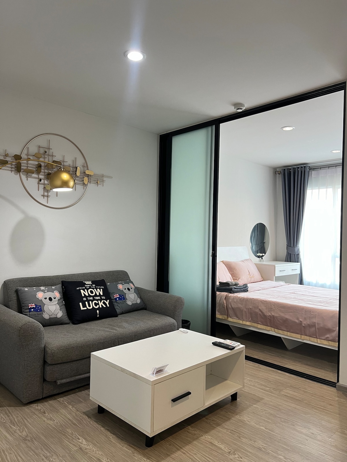 A cozy living area features a gray sofa with a decorative pillow, complemented by a coffee table. The space is divided by glass panels, revealing a bedroom area with a spacious bed dressed in soft linens. Natural light filters in through sheer curtains.