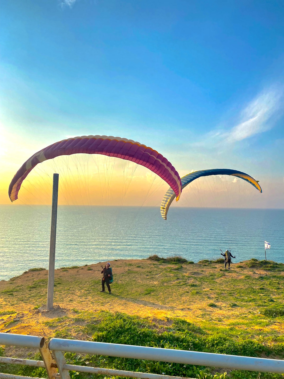 Two colorful paragliders are seen soaring above the coastline, framed by a pastel sky at sunset. A grassy area in the foreground leads towards the edge of the cliff, where a person is preparing for flight. The vast ocean stretches in the background.