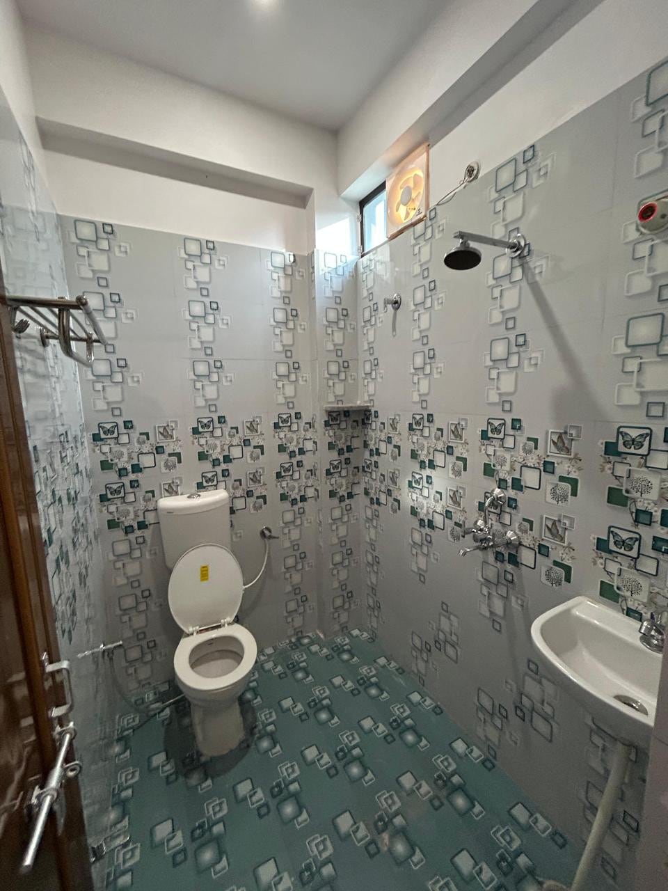 A modern bathroom is showcased, featuring a showerhead above a tiled wall. A toilet and a small sink are positioned against the patterned wall. The floor displays a blue-green tile design that complements the decor, while a small window provides natural light.