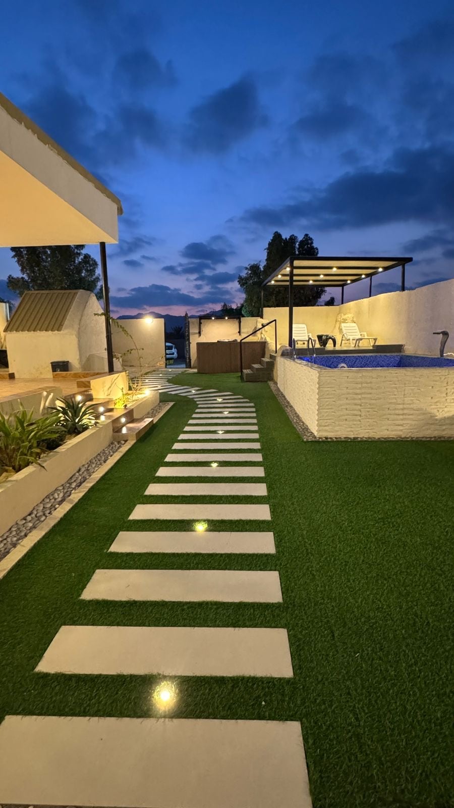 A well-maintained outdoor area features a pathway lined with rectangular stepping stones, leading towards a private swimming pool. Subtle lighting highlights the greenery and landscaping, while a shaded seating area is visible in the background, creating a serene atmosphere as twilight approaches.