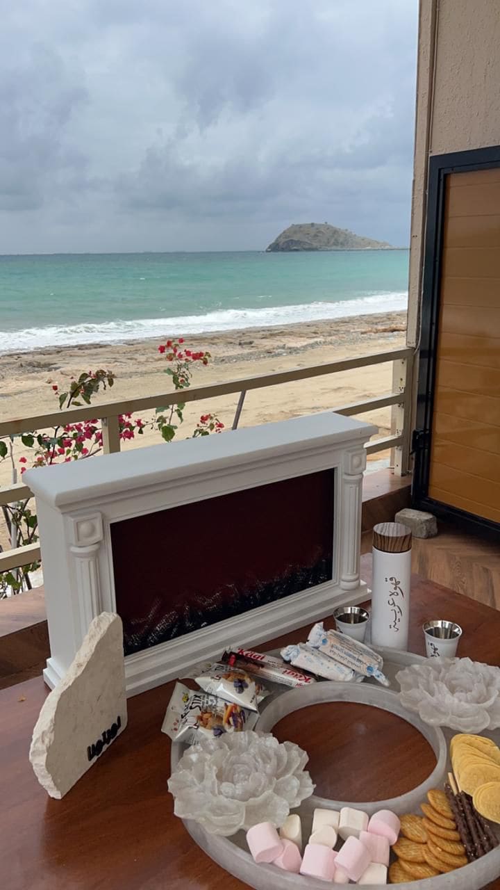 A decorative electric fireplace is positioned with a view of the beach and distant island. A variety of snacks, including marshmallows and cookies, are arranged on a circular tray. Flowering plants can be seen outside near the water's edge.