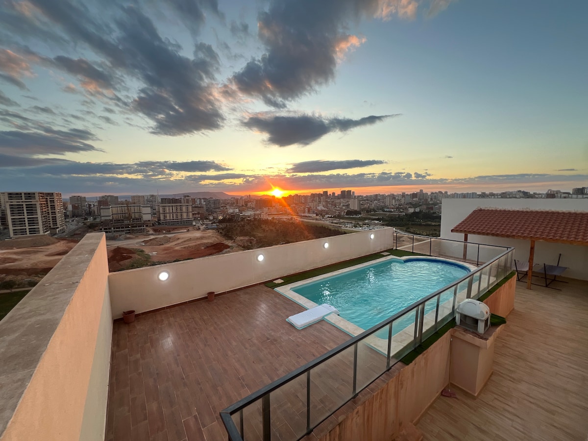 An outdoor terrace features a rectangular swimming pool surrounded by wooden decking. The sunset casts warm hues across the sky, framing the distant city skyline. Soft lighting enhances the area, and a covered seating area offers a place to relax.