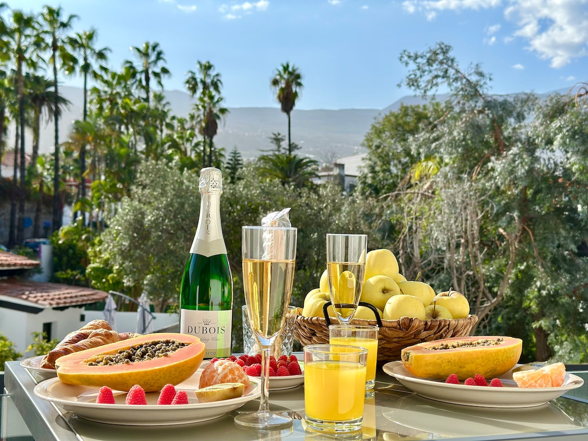 An outdoor dining setup is displayed on a terrace, featuring a variety of fruits, croissants, and sparkling beverages. Lush greenery and palm trees frame the background, while mountains are visible in the distance under a clear sky.