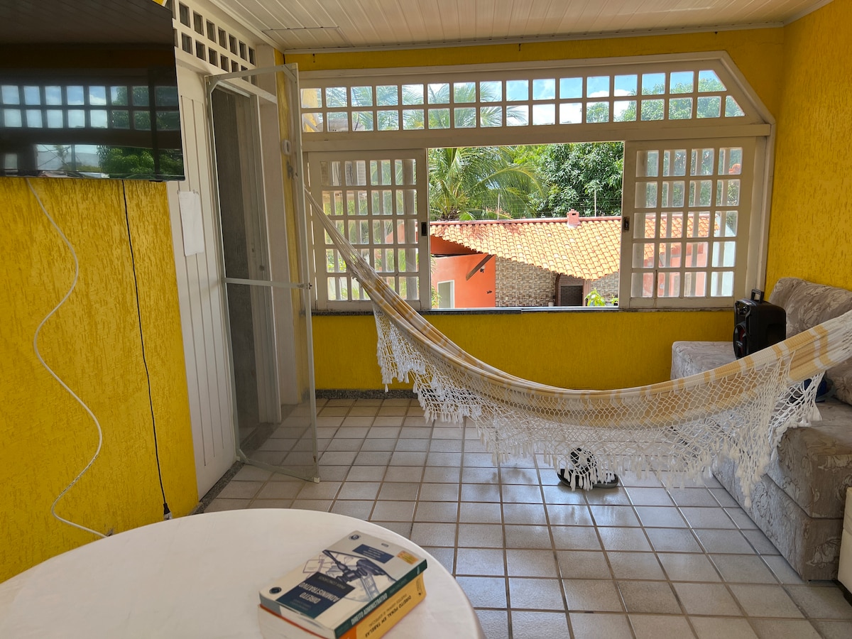 A bright and airy living area features large windows allowing natural light, complemented by vibrant yellow walls. A hammock is hung across the space, while a light-colored couch provides seating. A round table is visible in the foreground, adorned with a book.