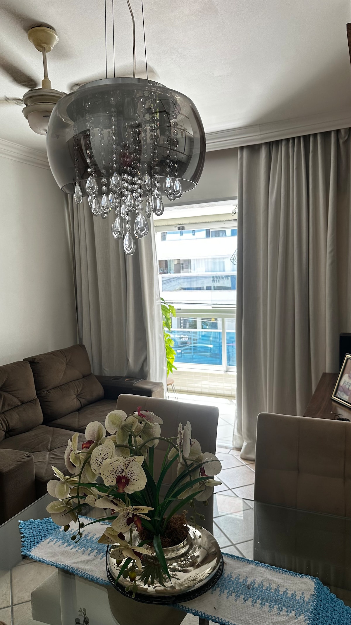 A stylish living space features a modern chandelier with hanging glass accents, suspended above a glass dining table. A decorative vase with orchids sits centrally, while natural light filters through a large window adorned with sheer curtains, revealing an outdoor view.