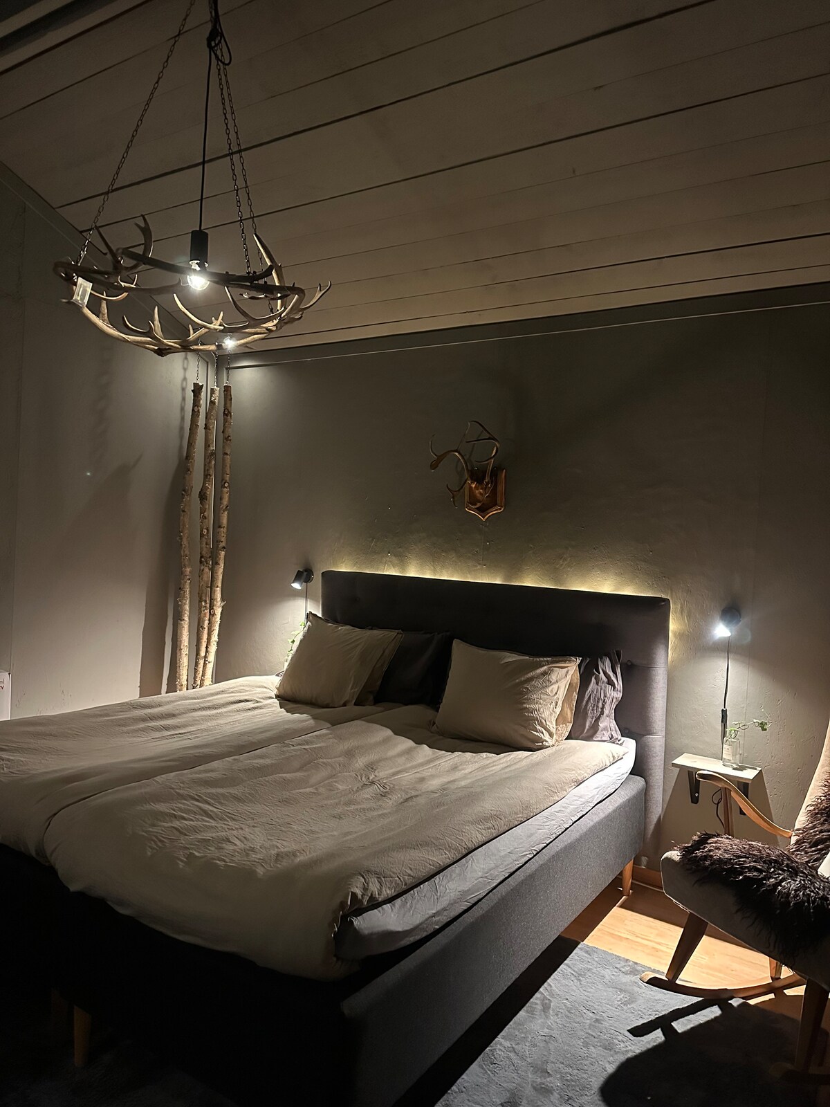 A cozy bedroom is featured, showcasing a spacious bed with neutral bedding. Soft lighting from wall-mounted lamps and a chandelier creates a serene atmosphere. Natural elements, such as a wooden sculpture and deer antlers, are included in the decor, enhancing the rustic charm.