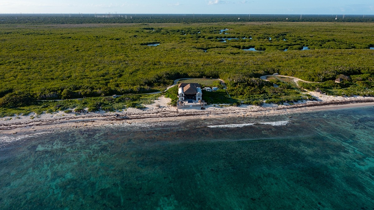 The image presents a serene beachfront property surrounded by lush greenery and natural landscapes. Clear blue waters gently lap at the sandy shore, while a house sits peacefully, offering direct access to the beach. The expansive view highlights the tranquility of the setting.
