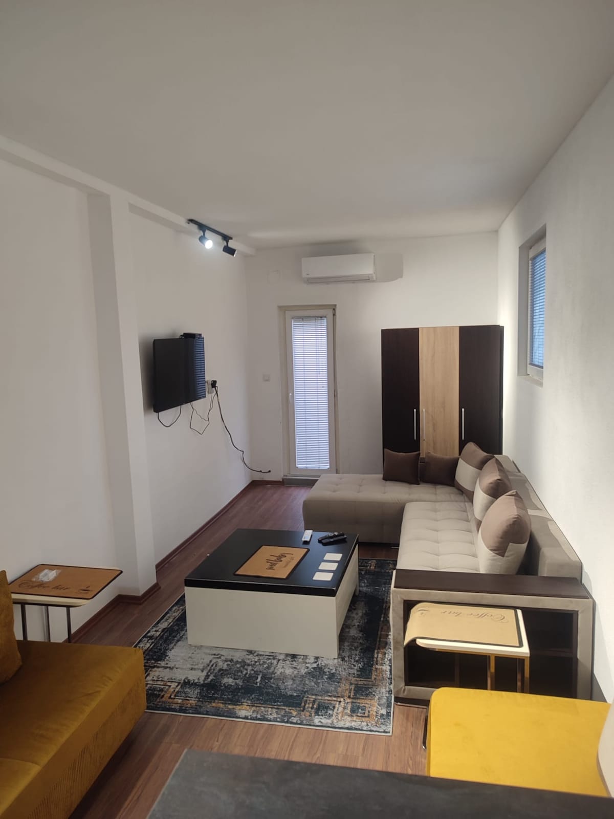 A bright living area is furnished with a modern sectional sofa and a coffee table centered on a patterned area rug. A wall-mounted TV is positioned opposite the seating, and a sliding door provides access to the exterior. Warm lighting enhances the comfortable environment.