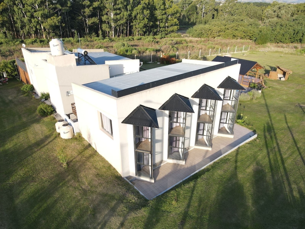 An aerial view showcases a modern white building with multiple protruding windowed structures. The surrounding landscape features lush greenery and a clear sky, emphasizing the serene and spacious outdoor area.