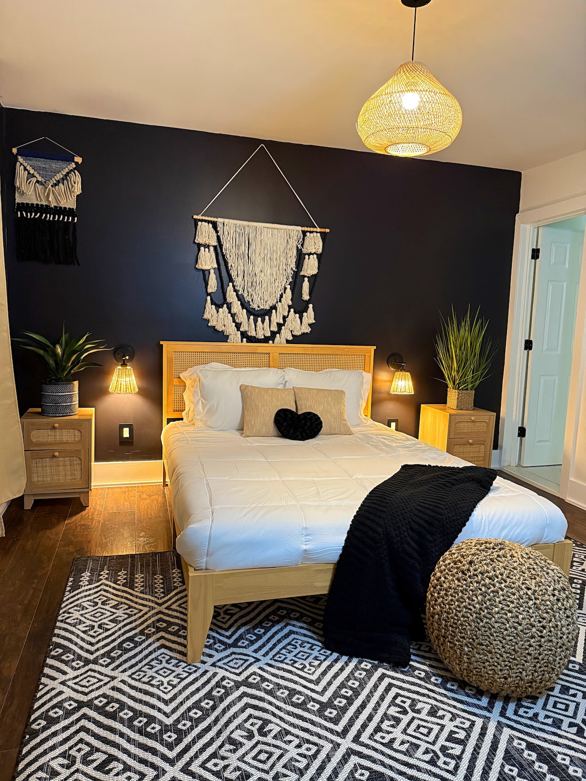 A cozy bedroom features a spacious bed with a white comforter and a heart-shaped pillow. The walls are painted dark blue, accented by wall hangings. Two bedside tables with lamps flank the bed, and a large area rug adds warmth. Indoor plants are positioned in the corners.