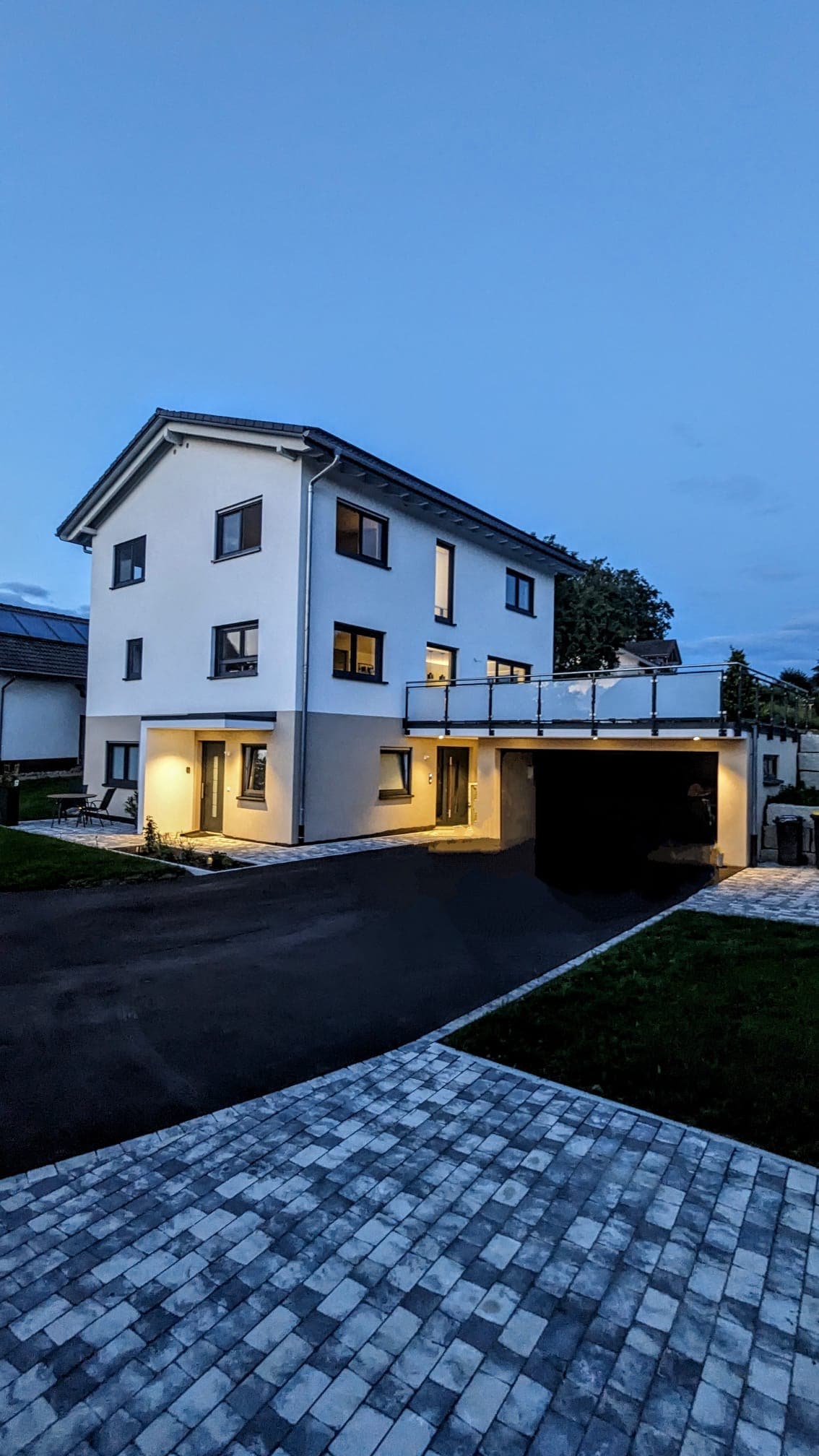 A modern, two-story building is shown in the evening light. The exterior features large windows, a flat roof, and a garage. Soft illumination highlights the entrance and a terrace above, while a neatly paved driveway enhances the approach to the property.