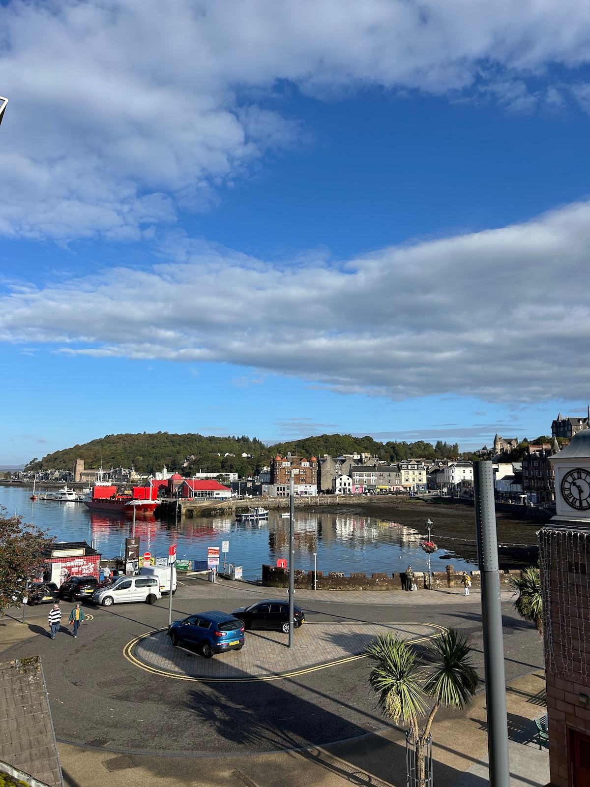 A waterfront view captures the serene harbor, featuring calm waters reflecting a vibrant blue sky. Various boats are docked, with colorful buildings lining the shore. Palm trees provide a touch of greenery, while a road in the foreground indicates accessibility to the area.