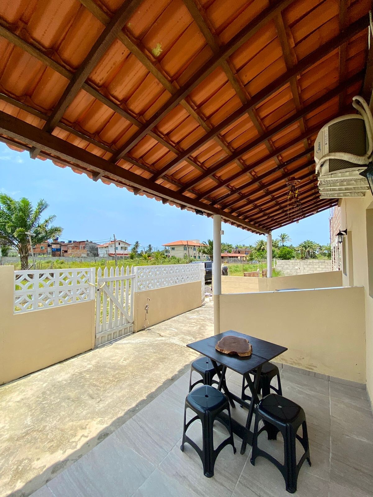 A covered porch area features a small black table accompanied by three matching chairs. A portion of the driveway and a gate are visible in the background, with greenery and distant buildings creating a serene outdoor atmosphere.