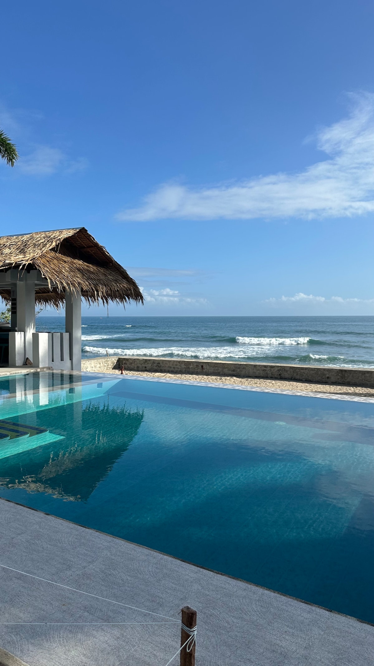 An infinity pool reflects the clear sky and ocean waves, bordered by a sandy beach. A thatched-roof structure provides shaded seating, enhancing the beachfront view. Gentle waves can be seen lapping at the shore, contributing to the serene atmosphere of the location.