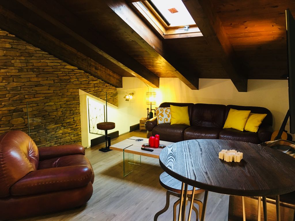 The lofted living area features wooden beams and a stone wall, creating a cozy atmosphere. Two leather sofas, one with yellow cushions, are arranged around a coffee table. A dining table is positioned nearby, and natural light enters through a skylight above.