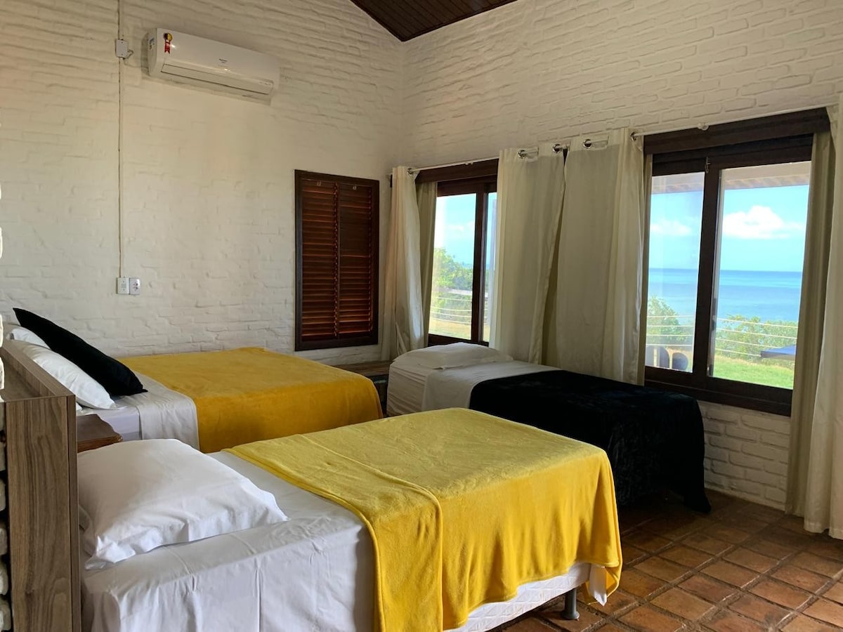 A bright sleeping area showcases three beds, including a spacious double bed, a single bed, and another single bed. Large windows provide a panoramic view of the ocean, while soft light illuminates the room. Light-colored walls complement the terracotta floor.