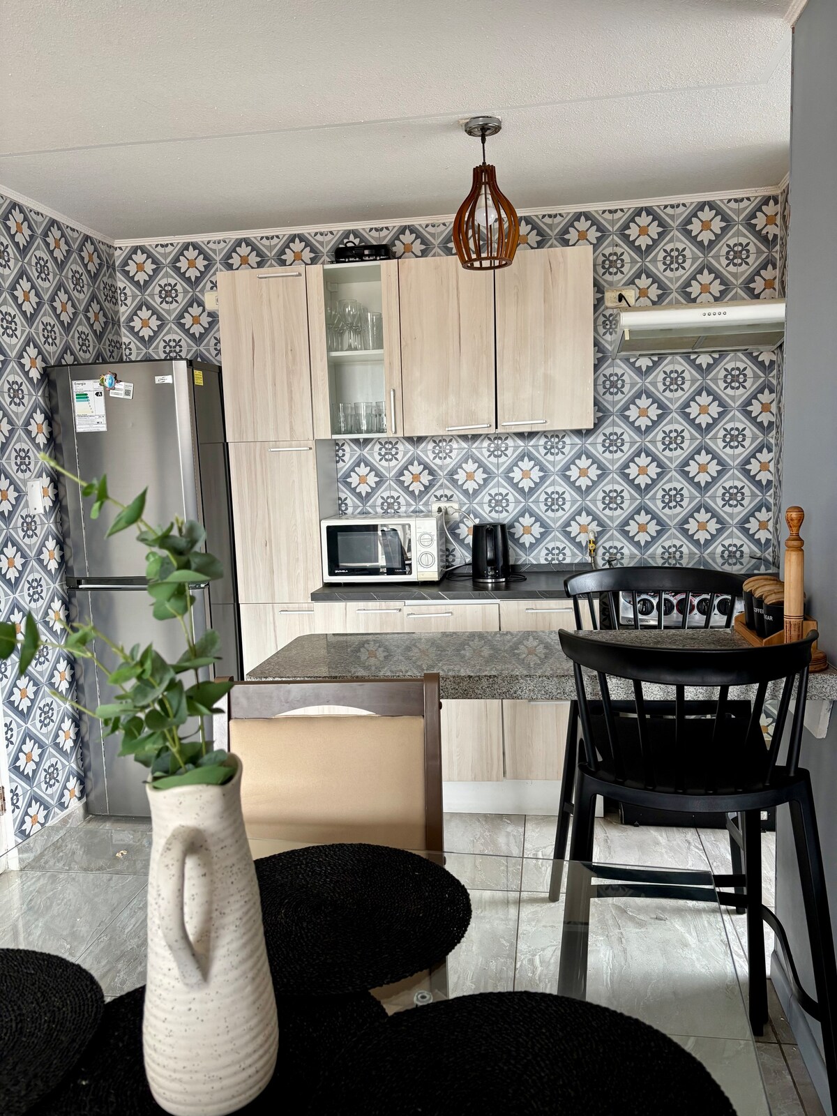 The kitchen features modern cabinetry and a patterned wall, creating a fresh ambiance. A dining table is complemented by a black chair, with a vase on the table. Essential appliances, including a microwave and coffee maker, are arranged neatly for convenience.