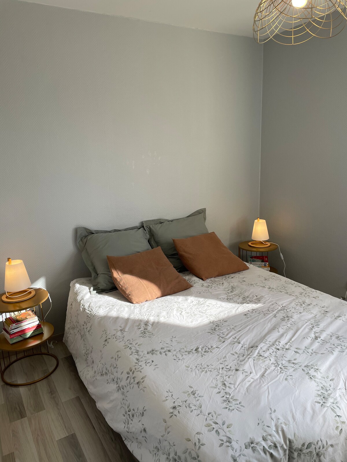 A cozy bedroom features a spacious bed adorned with a floral-patterned bedspread and decorative pillows. Bedside tables with gentle lighting are positioned on either side, while a stack of books sits neatly beside one lamp. Soft grey walls provide a calming backdrop.