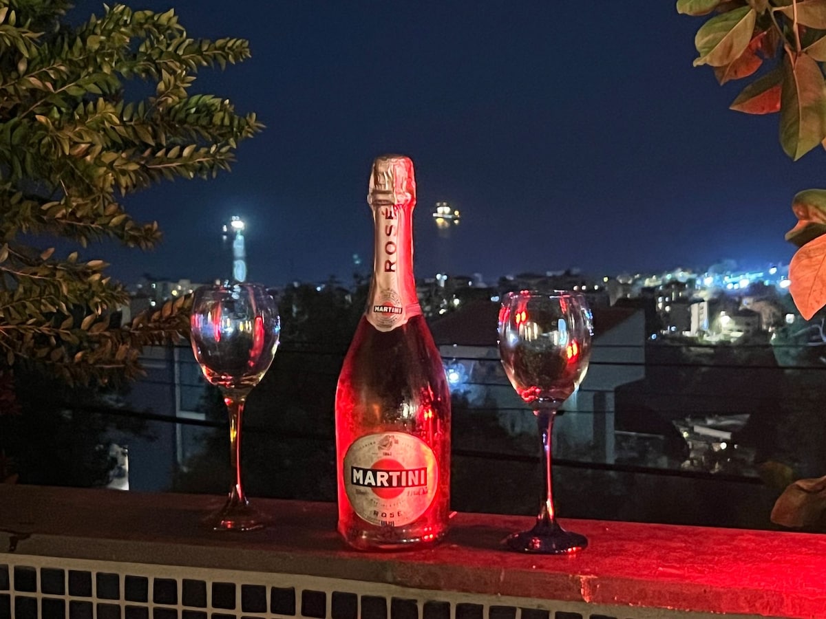 A bottle of Martini Rosé stands on a ledge with two elegant glasses nearby. The scene is illuminated by soft red lighting, while a cityscape with twinkling lights is visible in the background, creating an inviting evening atmosphere.
