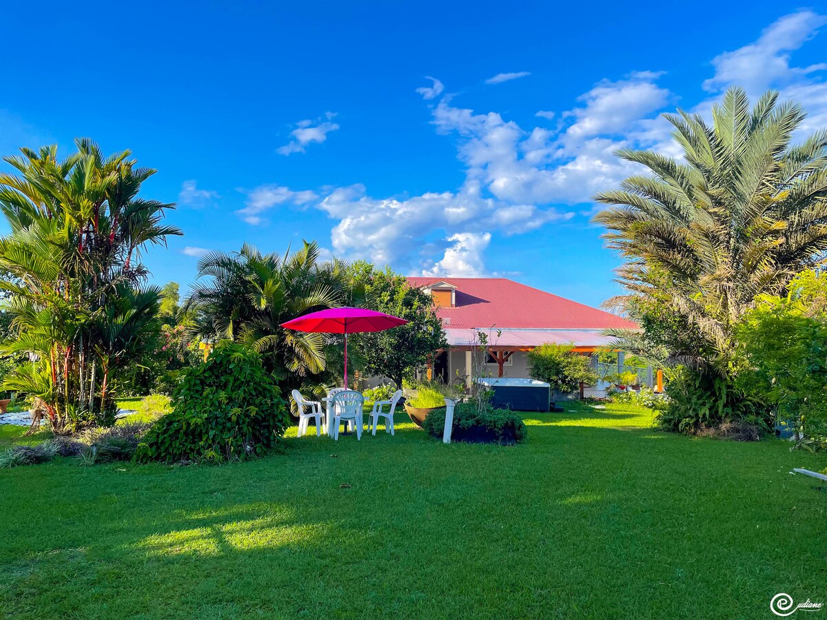 A spacious villa is surrounded by lush greenery and palm trees. A red umbrella shades a seating area with white chairs, placed on well-maintained grass. The house features a wide veranda and a contrasting red roof, offering an inviting outdoor space.