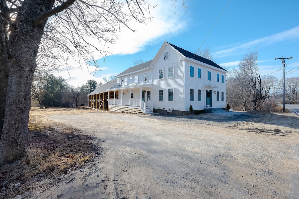 A two-story white farmhouse is presented with a symmetrical design, featuring multiple windows and a large covered porch. The surrounding area includes a gravel driveway and sparse trees, contributing to the property's spacious outdoor setting. A clear blue sky enhances the peaceful atmosphere.