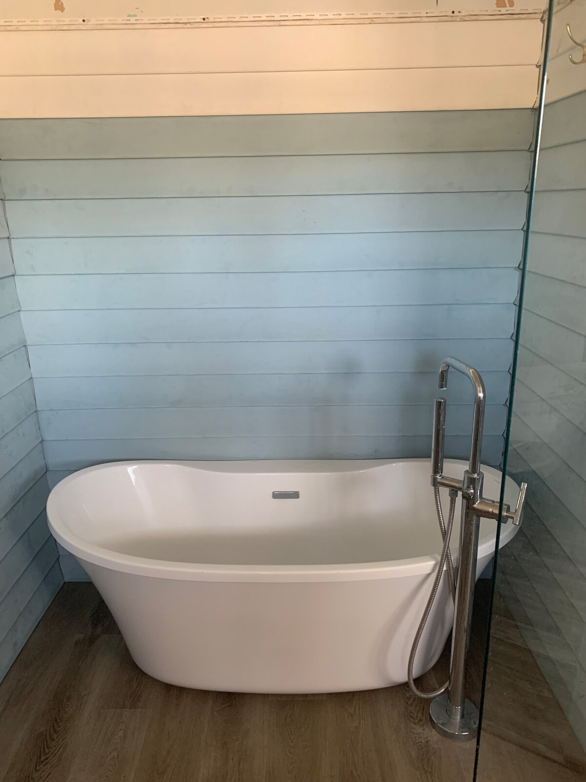 A freestanding bathtub is positioned in a serene bathroom featuring light blue shiplap walls. A sleek chrome shower fixture is attached nearby, with a clear glass panel providing separation while maintaining an open feel. Natural light may enhance the calm atmosphere in this tranquil space.