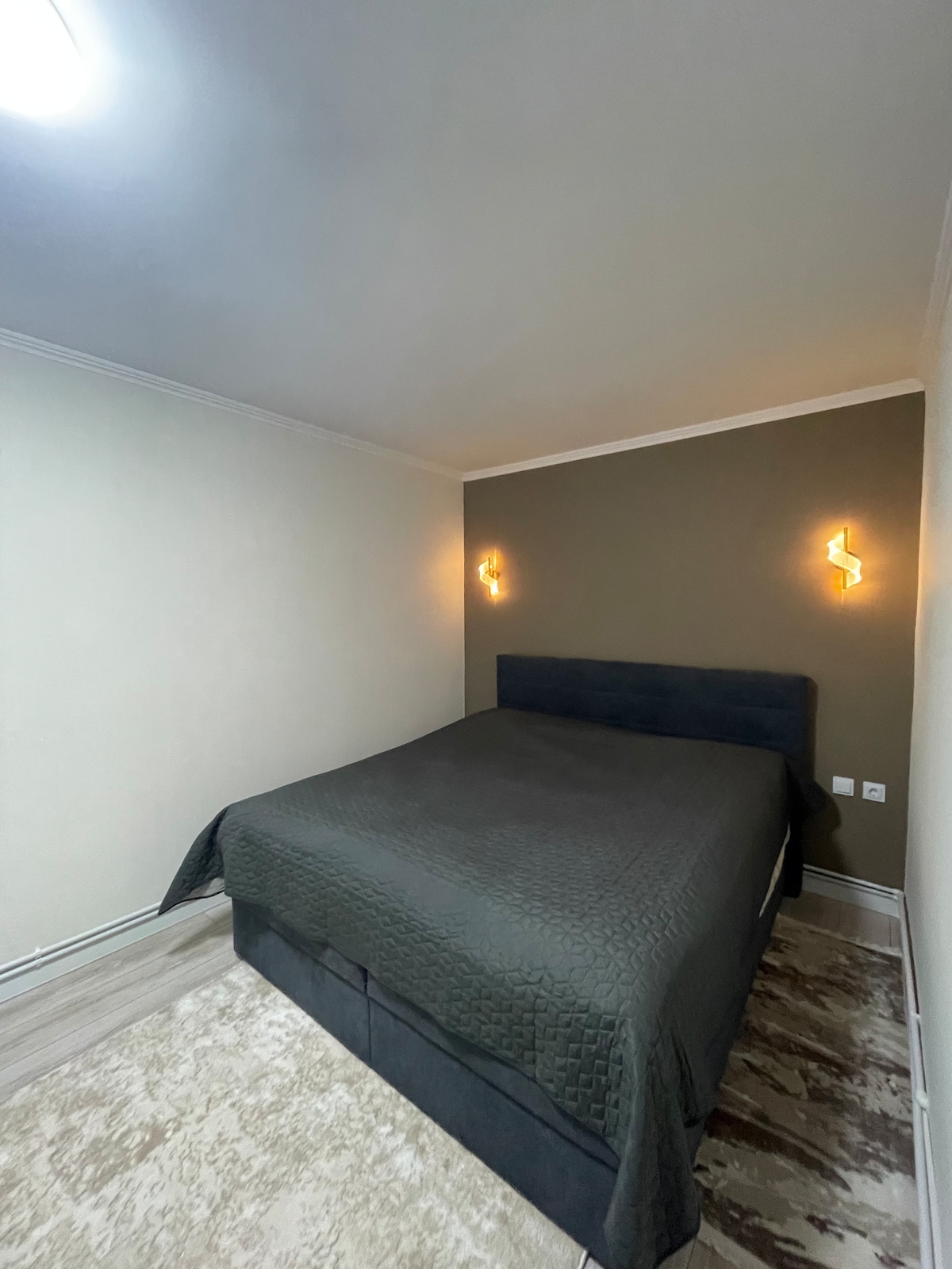 A cozy sleeping area is highlighted by a spacious bed dressed in a dark quilt. Soft lighting is provided by wall sconces on either side, while neutral walls and a patterned rug contribute to a serene atmosphere.