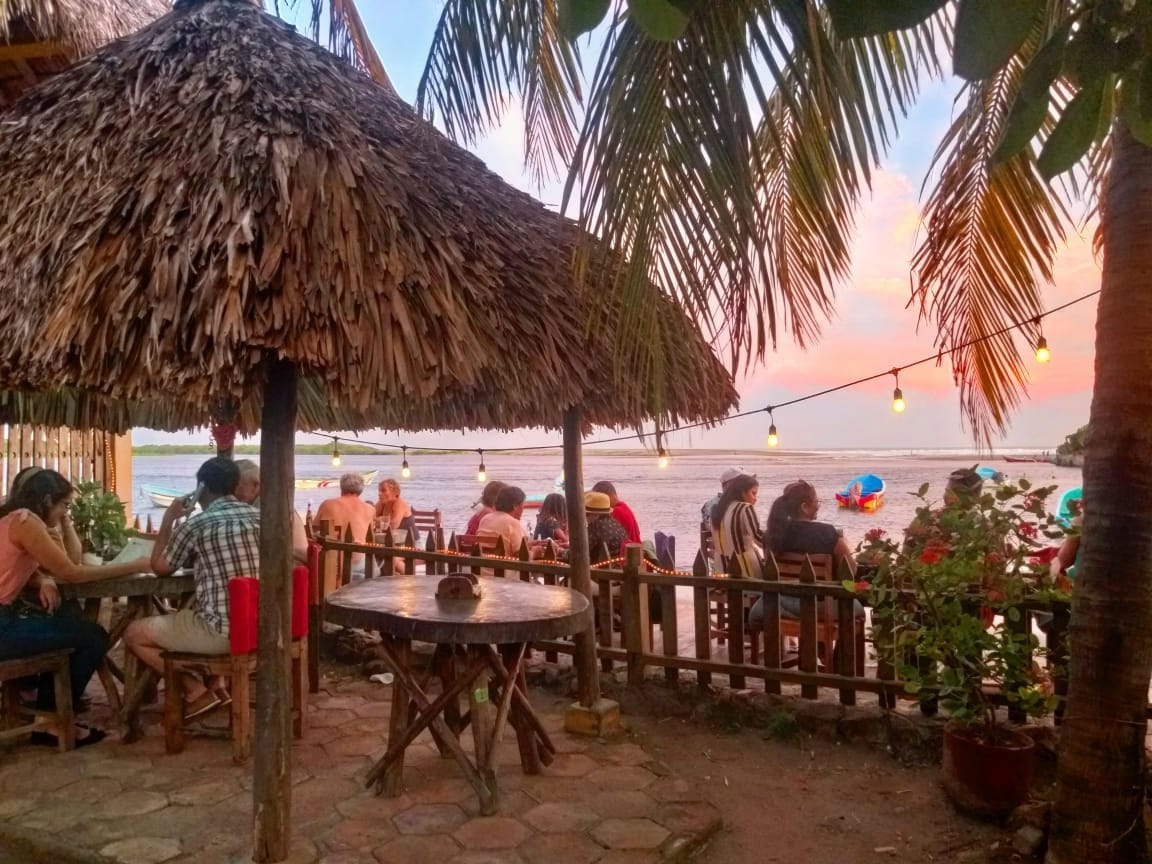 A casual outdoor dining area features thatched-roof tables surrounded by palm trees. Guests enjoy their meals while colorful kayaks are visible on the water at sunset. Strings of lights add to the ambiance, illuminating the atmosphere as the sky transitions to soft hues.