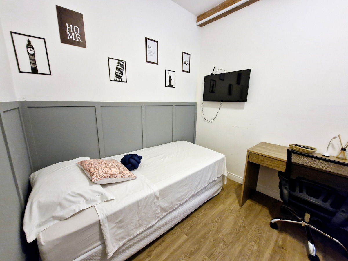 A single bed is positioned against a grey-paneled wall, featuring decorative black-and-white wall art. A desk with a chair is situated nearby, with a television mounted on the opposite wall. Natural light illuminates the wooden flooring.