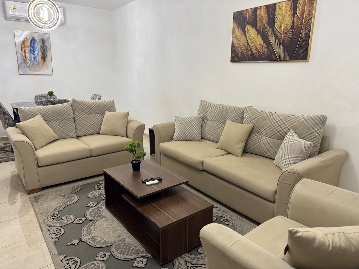 A comfortable seating area features two light beige sofas adorned with patterned throw pillows. A dark wooden coffee table sits in the center, complemented by a small potted plant. A decorative wall art piece depicting feathers adds a subtle touch to the setting.