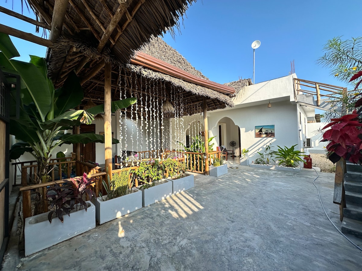 An outdoor area features a thatched roof and decorative elements, including hanging strings of lights. Lush greenery and various plants are arranged around white planter boxes. Sunlight casts soft shadows on the concrete surface, creating a bright and inviting atmosphere.