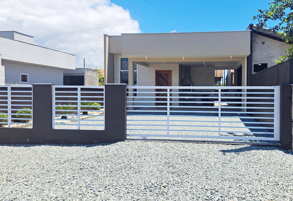 The exterior of the property is showcased, featuring a modern structure set behind a sleek white gate. Gravel surrounds the entrance, leading to a covered area with a barbecue setup. The design emphasizes clean lines and a welcoming entrance, bordered by minimalist landscaping.