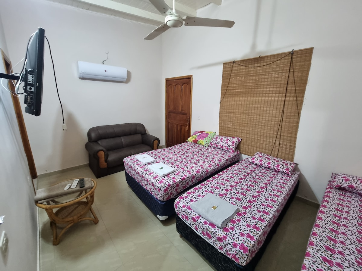 A cozy bedroom features three beds with floral-patterned linens. A leather sofa is positioned against the wall, with a wooden table nearby. A television hangs on the wall opposite the beds. Natural light is filtered through bamboo curtains, creating a comfortable ambiance.