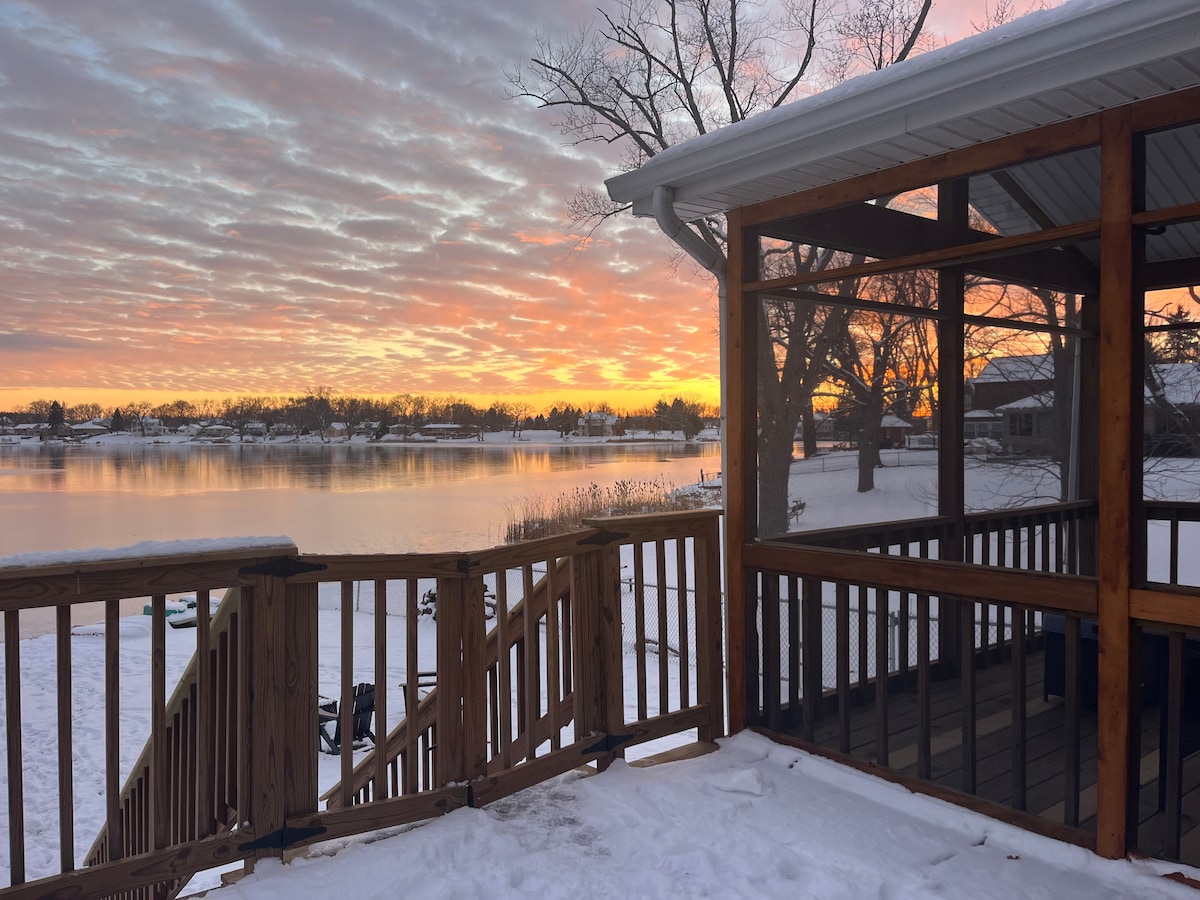 A serene lakeside view is presented at sunset, with colorful clouds reflecting on the frozen surface of the lake. The image captures the wooden deck leading to the screen room, surrounded by snow. Views extend to the opposite shore, revealing a landscape of trees and homes.