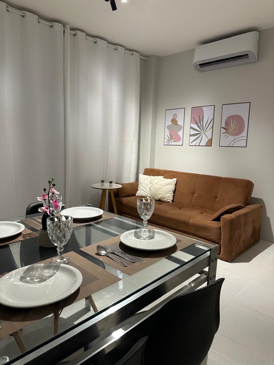 A dining area features a glass table set for four with plates and cutlery. A brown sofa is positioned against the wall, complemented by framed botanical artwork. Sheer curtains allow natural light to enter, while an air-conditioning unit is visible above the sofa.