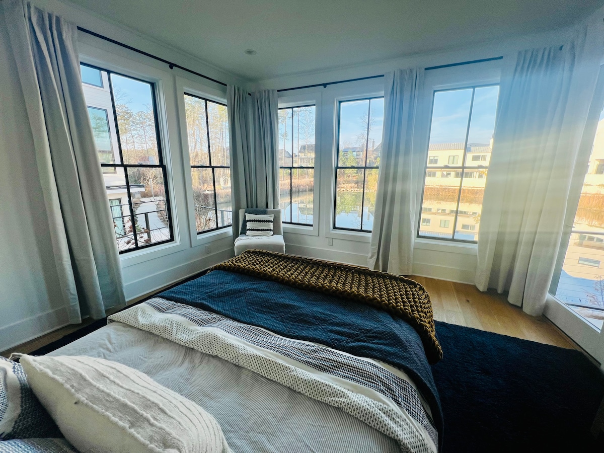 A cozy bedroom features large windows with soft curtains, allowing natural light to illuminate the space. A spacious bed is adorned with a textured blanket, and a light accent chair is positioned nearby, offering a view of the serene surroundings outside.