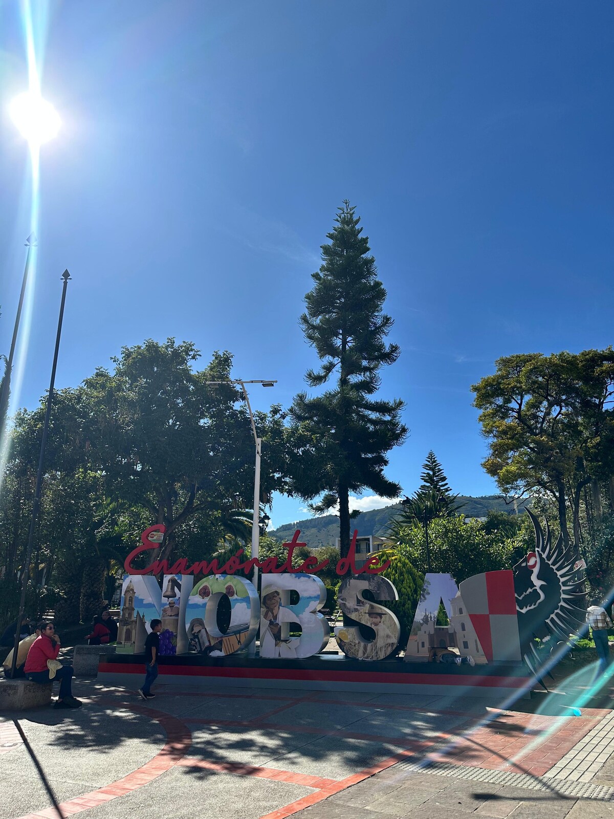 The image features a large, colorful sign reading 'NOBSA' prominently displayed in an outdoor area. Surrounding trees provide greenery, while a clear blue sky enhances the inviting atmosphere. People can be seen nearby, creating a lively community environment.