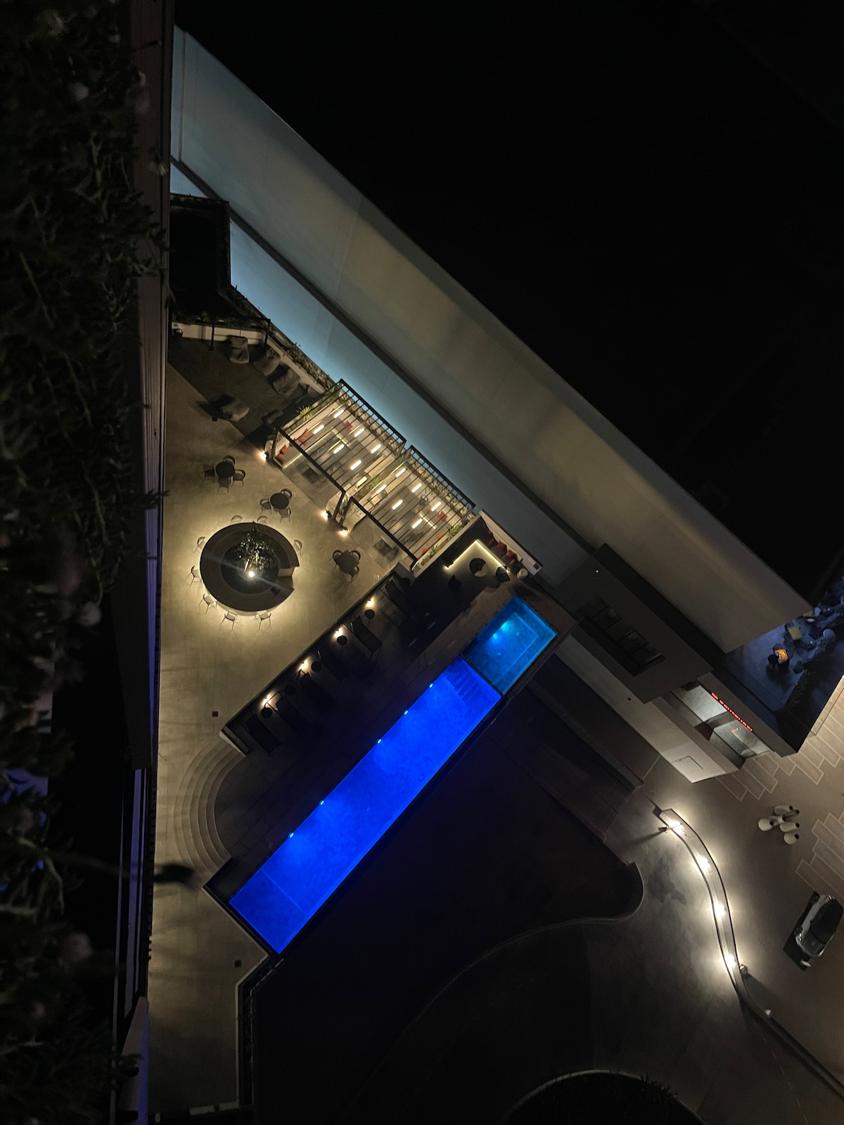 An aerial view captures the outdoor space featuring a long, illuminated rectangular pool and a circular seating area with a fire pit. Soft lighting highlights the modern design of the patio and poolside, creating an inviting atmosphere for relaxation.