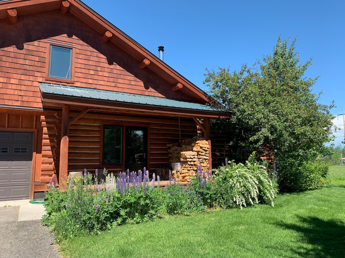 The exterior of a log cabin features a welcoming entrance with wooden beams and a sloped roof. Colorful flowers and lush greenery surround the front, providing a vibrant contrast to the earthy tones of the logs. A driveway and garage door are visible on one side.